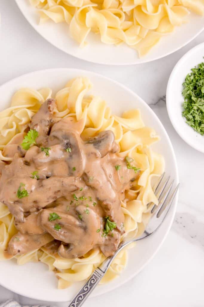 Beef stroganoff served over a bed of egg noodles on a white plate with a fork. Parsley garnish on top.