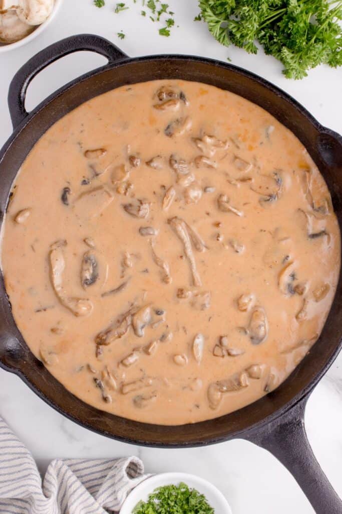 Completed beef stroganoff recipe in a cast iron pan on a table surrounded by mushrooms, parsley, and a towel.