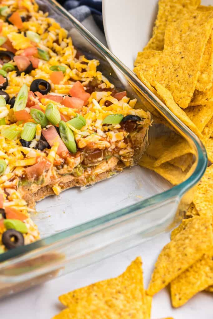 Glass serving dish with a cut away of the 7 layer dip. Serving dish is surrounded with corn tortilla chips.