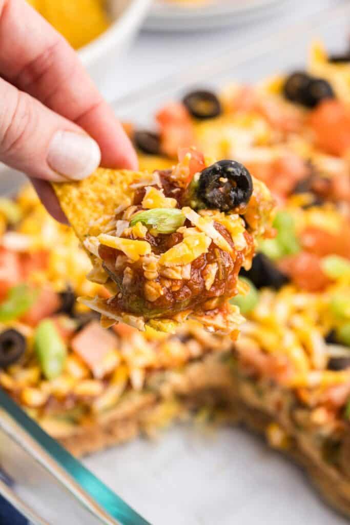 7 layer dip shown as a cut away in the background. The foreground shows a hand holding a corn tortilla chip with 7 layer dip on it.