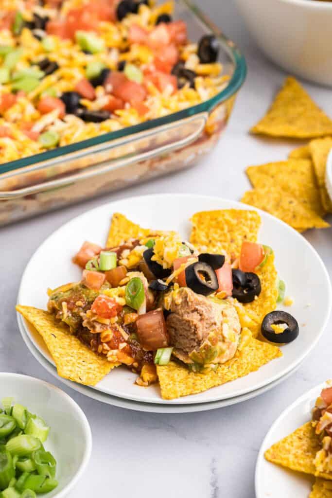 A white bowl of corn tortilla chips with 7 layer dip on top. Serving dish of 7 layer dip in the background.