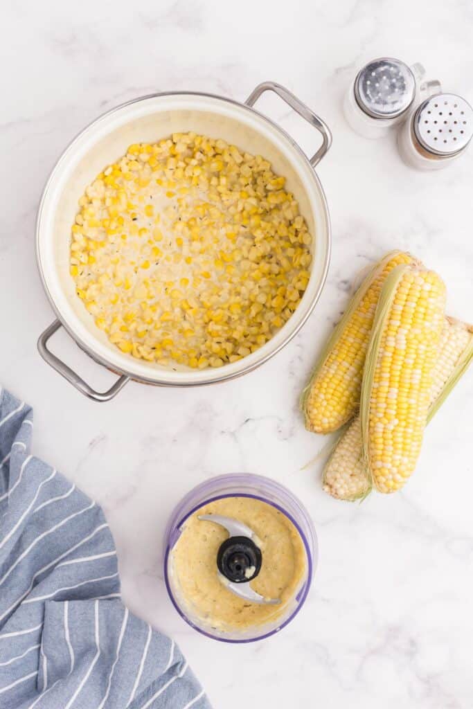 Pot of creamed corn surrounded by salt, pepper, corn on cob, and a small food processor with corn mixture in it.