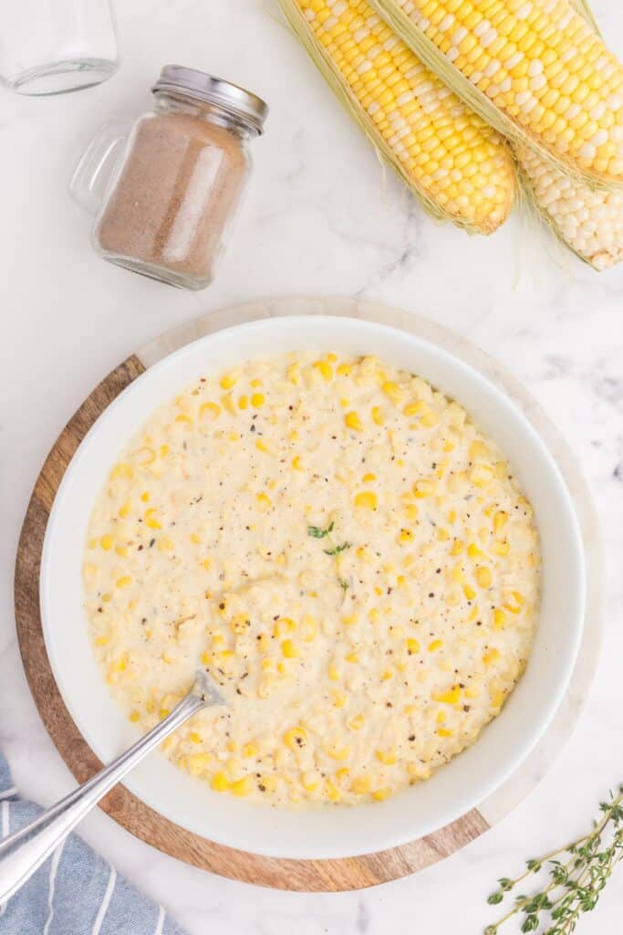 Overhead image of creamed corn in a serving bowl with a silver serving spoon. Surrounded by salt and pepper shakers, and several ears of corn on the cob.