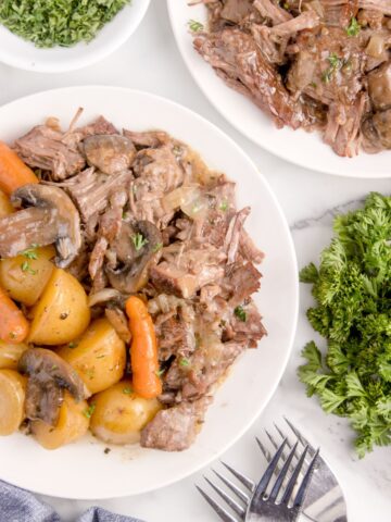 overhead view of 2 plates with chuck roast with carrots, potatoes, and mushrooms garnished with parsley.