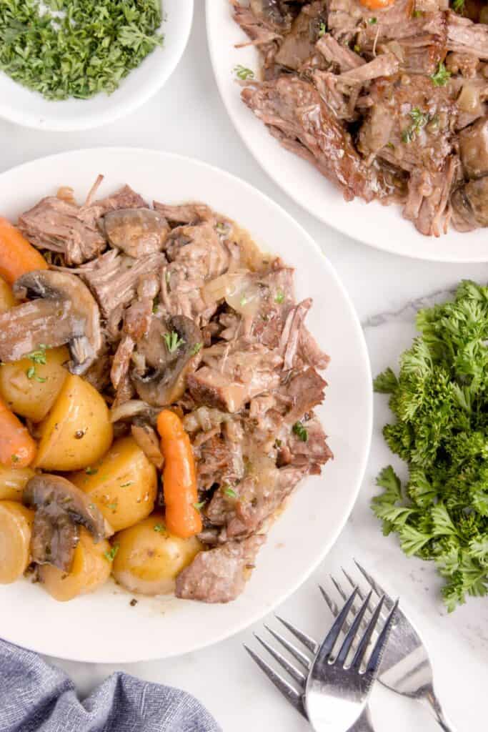 overhead view of 2 plates with chuck roast with carrots, potatoes, and mushrooms garnished with parsley.