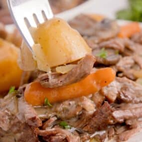 Fork full of potato, carrot, and chuck roast above a white plate.