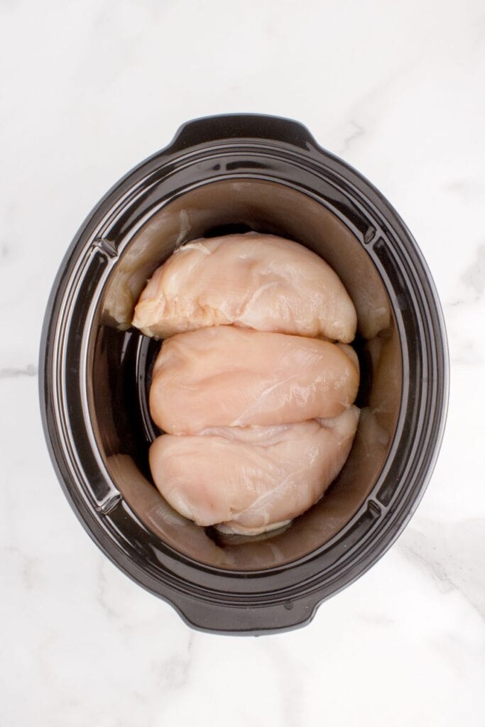 Raw chicken breast arranged at the bottom of the slow cooker pot.