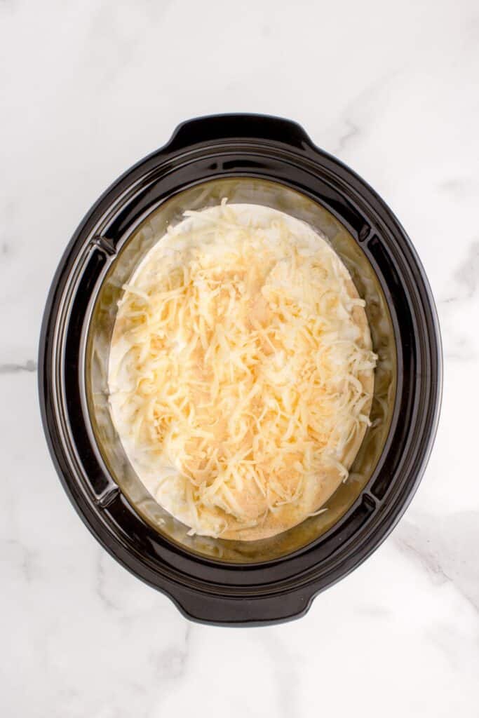 Overhead view of crock pot with chicken, parmesan sauce, heavy cream, onion powder, and parmesan layered on top.