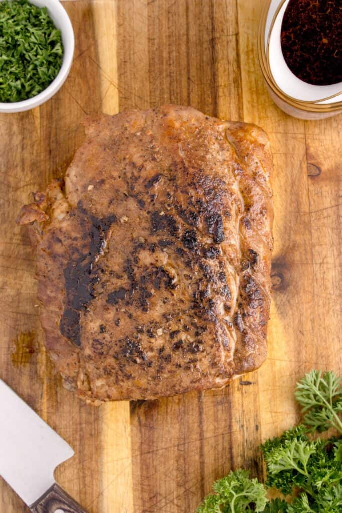 Completed pork tenderloin on a large cutting board. Surrounded by gravy in a gravyboat, parsley garnish, and a carving knife.