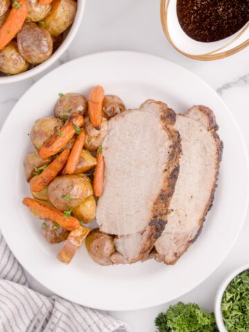 White dinner plate of sliced pork loin, potatoes, and carrots. Plate is surrounded by a bowl of parsley garnish, gravy in a gravy boat, and a bowl of potatoes and carrots.