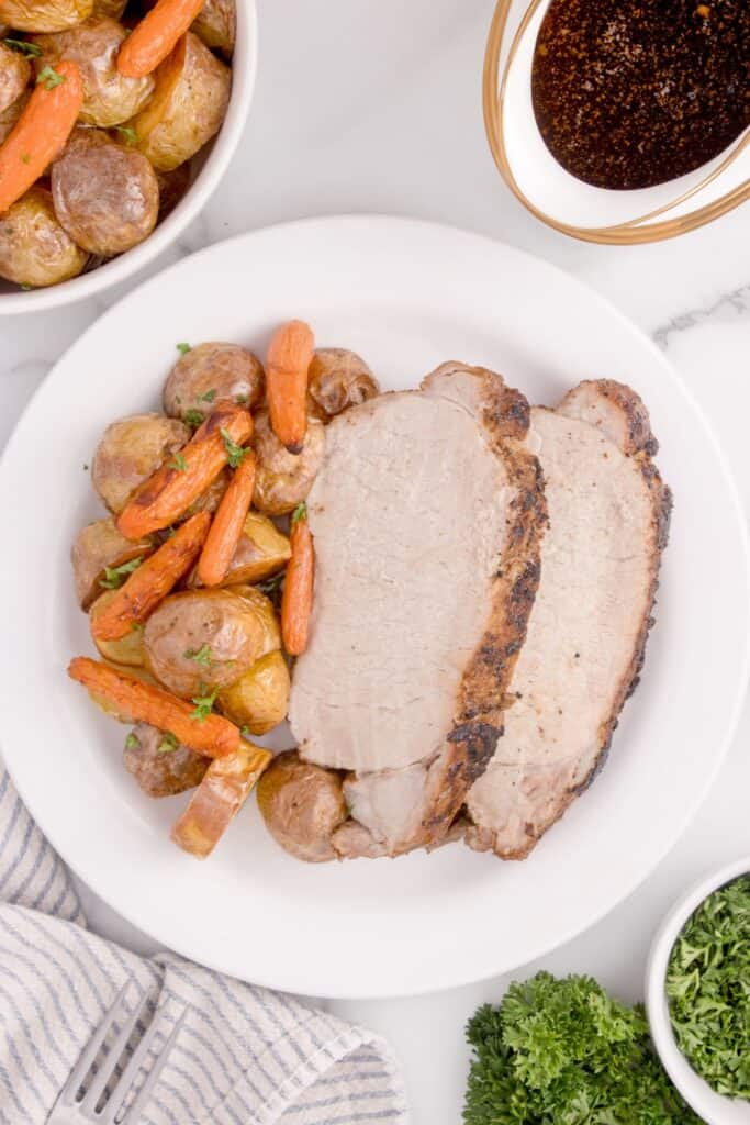 White dinner plate of sliced pork loin, potatoes, and carrots. Plate is surrounded by a bowl of parsley garnish, gravy in a gravy boat, and a bowl of potatoes and carrots.