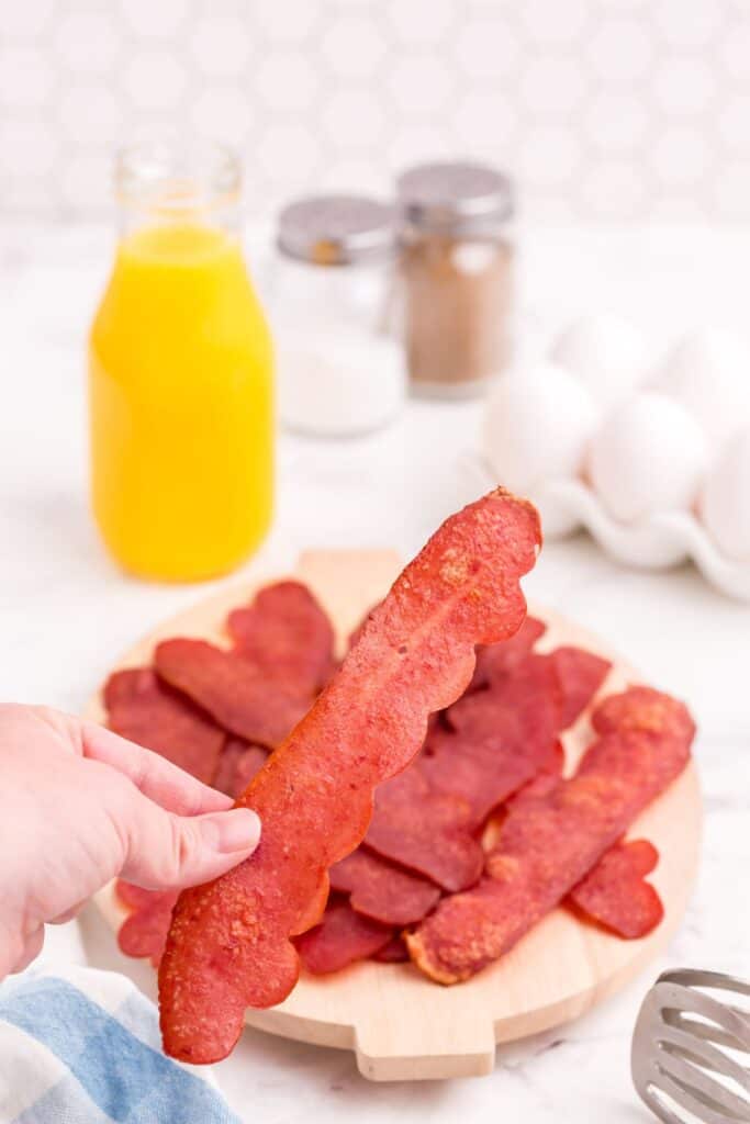 A hand is holding a single piece of oven baked turkey bacon in focus. In the background is a pile of turkey bacon on a cutting board, whole eggs, orange juice, and salt and pepper.