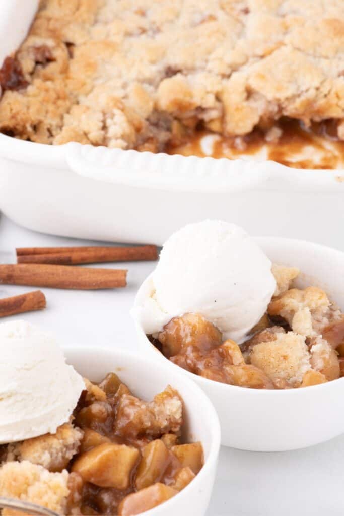 Two bowls of apple cobbler with ice cream in front of a baking dish of completed apple cobbler.