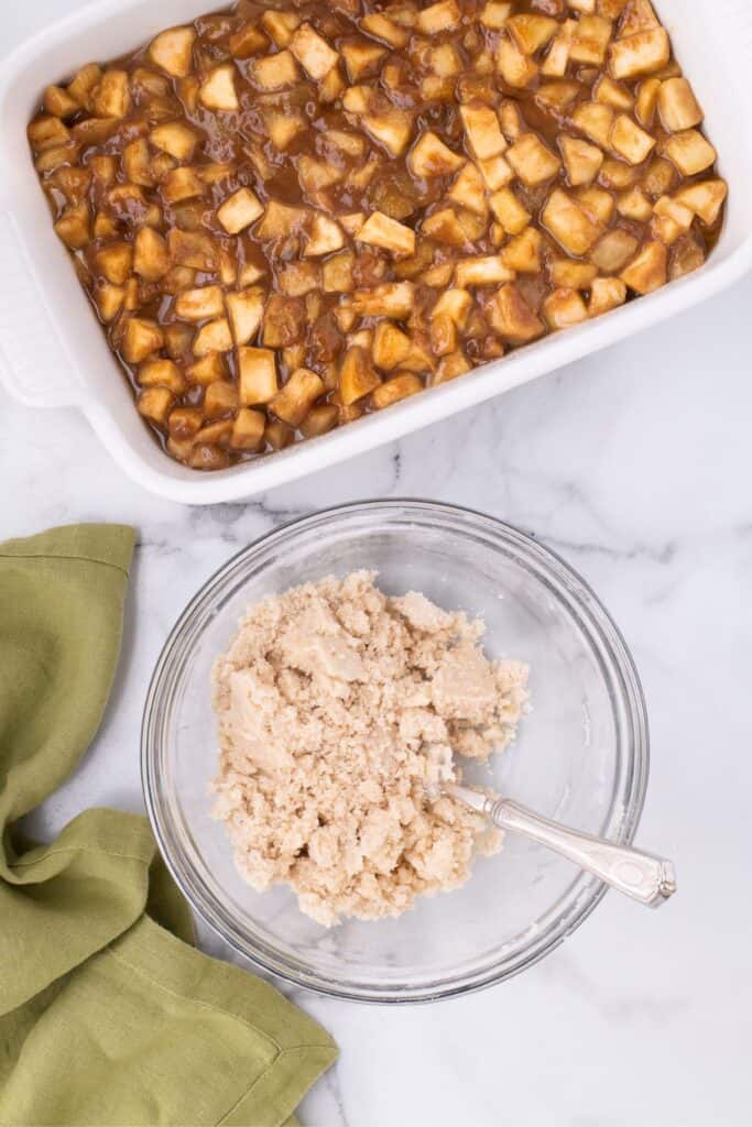 Crumble topping for apple cobbler in a glass bowl with a spoon next to baked apples in a baking dish.