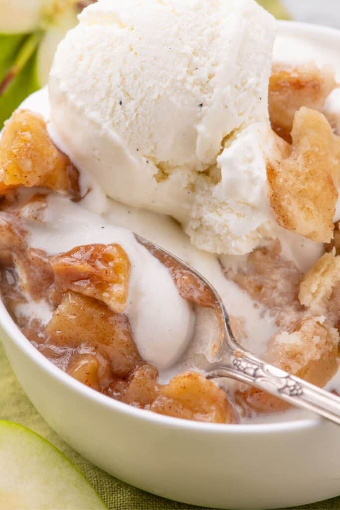 Close up image of apple cobbler with vanilla ice cream on top. Silver spoon scooping a bite.