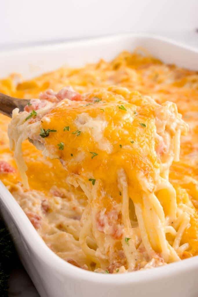 Wooden spoon lifting a serving of chicken tetrazzini out of a white baking dish.