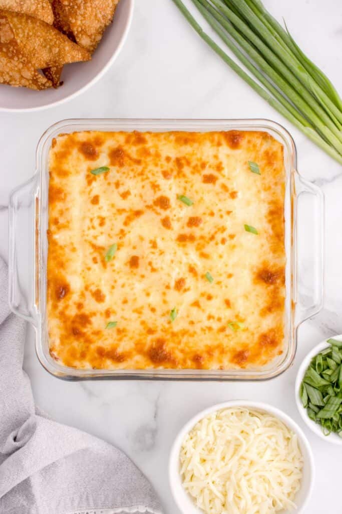 crab rangoon dip in a baking dish covered with mozzarella cheese, after baking. Dish is surrounded but whole green onions, wonton chips, a bowl of mozzarella cheese, and a bowl of chopped green onions.