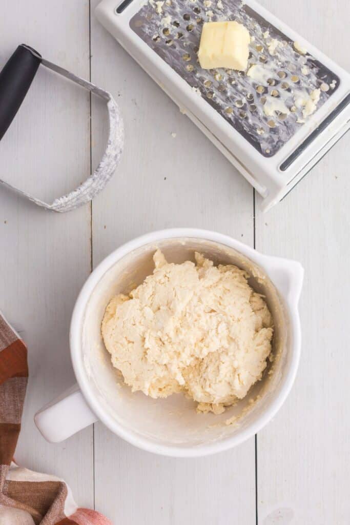 Biscuit dough in a bowl with a cheese grater and pastry cutter above it. Cheese grater is grating butter.