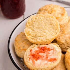 Serving bowl with biscuits piled up. Top biscuit is halved and spread with butter and strawberry jam.