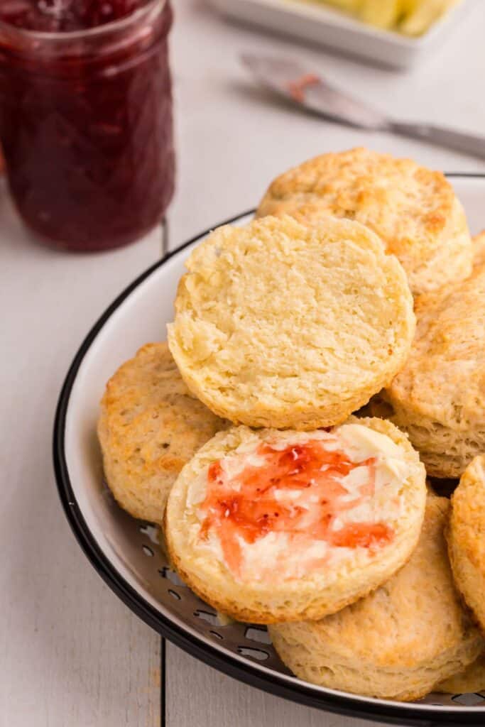 Serving bowl with biscuits piled up. Top biscuit is halved and spread with butter and strawberry jam.