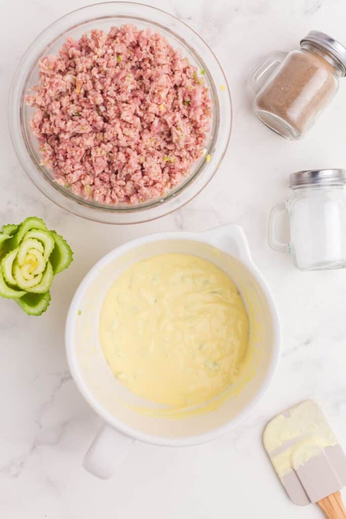 A white bowl of mayo mixture next to. glass bowl of chopped ham mixture.