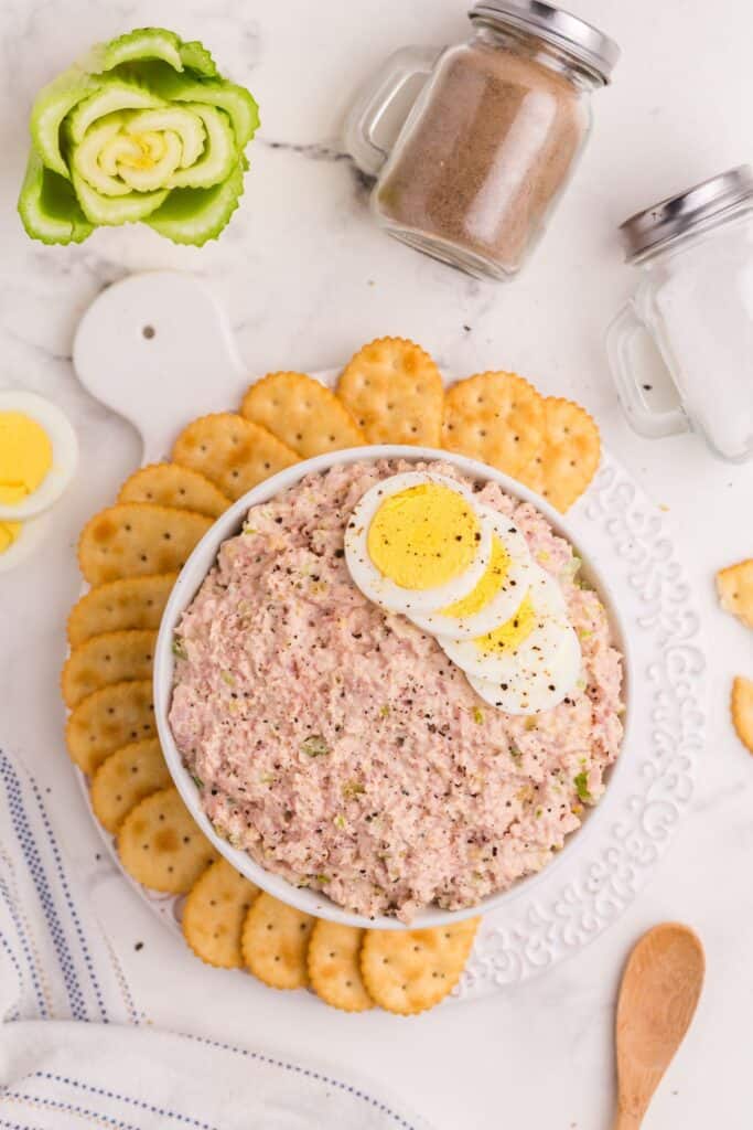 Overhead image of ham salad in a bowl. Bowl is surrounded by crackers, a wooden spoon, salt and pepper shakers.