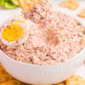 Ham salad in a bowl surrounded by crackers. A hand is scooping up ham salad with a cracker.