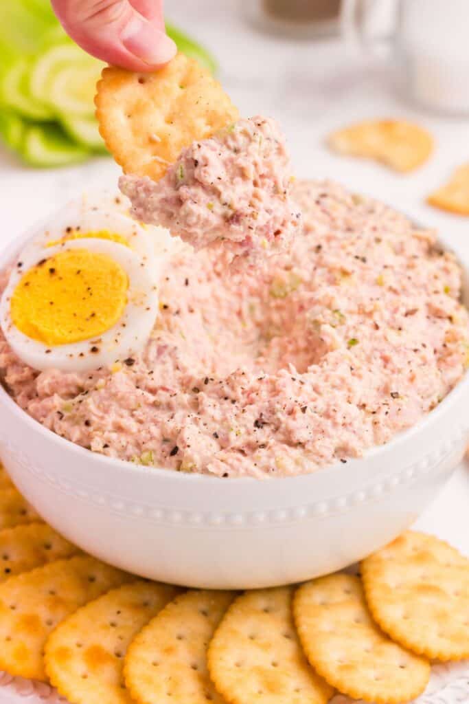 Ham salad in a bowl surrounded by crackers. A hand is scooping up ham salad with a cracker.