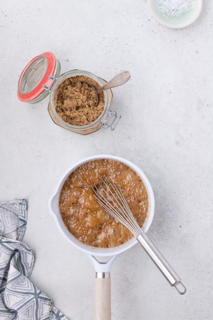 A brown sugar mixture is bubbling in a small saucepan. A jar of brown sugar and a bowl of salt are above the saucepan.