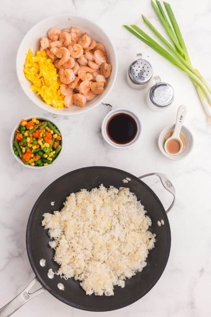 Shrimp fried rice ingredients around a skillet with white rice in it.