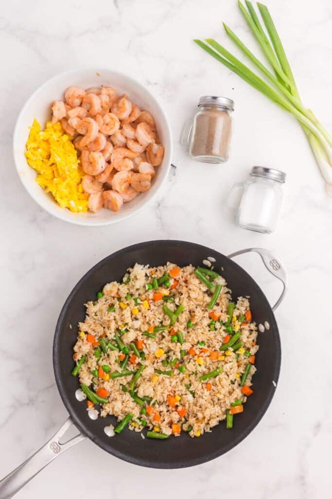 A bowl of cooked shrimp and scrambled eggs above a skillet with fried rice ingredients in it.