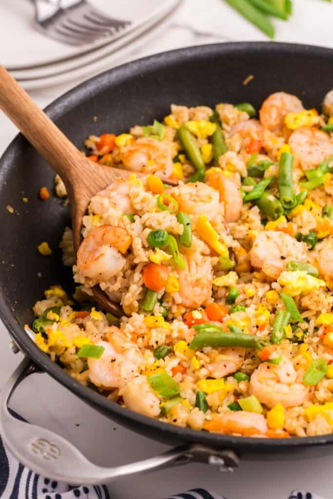 Close up view of a skillet with shrimp fried rice. A wooden spoon is stirring the fried rice recipe.