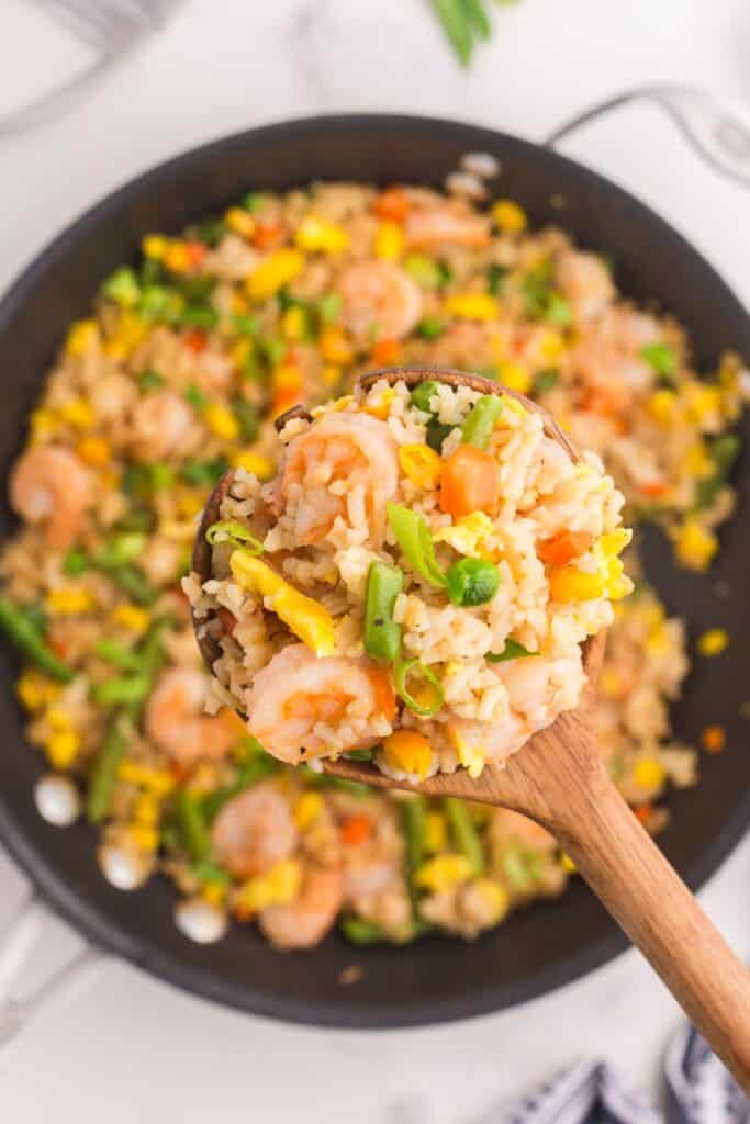 A wooden spoon is lifting a serving of shrimp fried rice above the skillet.