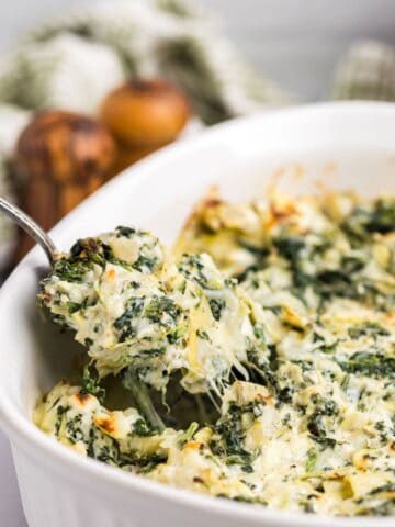 Spinach and artichoke dip in a white serving bowl with a spoon lifting out a serving of dip.