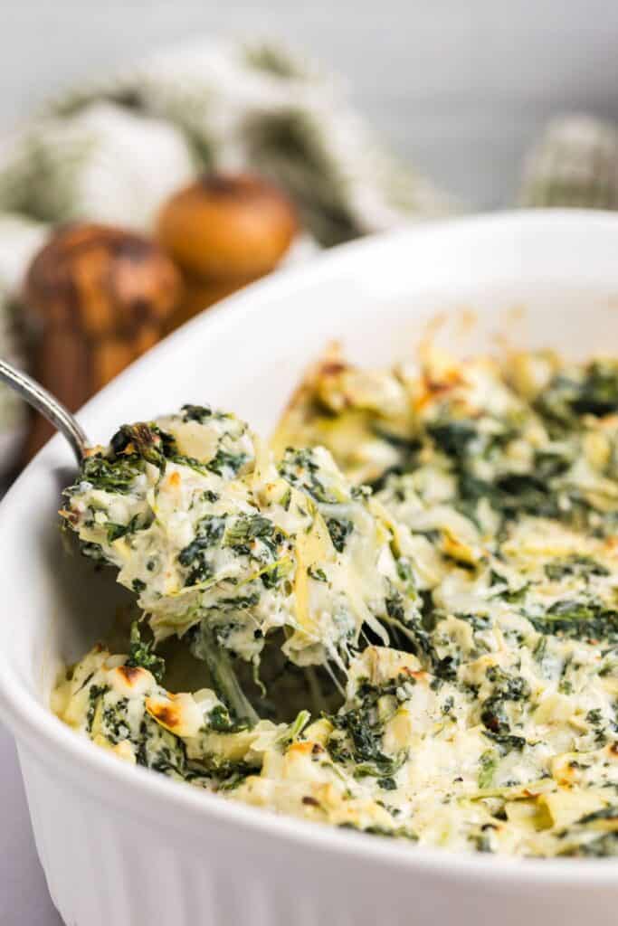 Spinach and artichoke dip in a white serving bowl with a spoon lifting out a serving of dip.