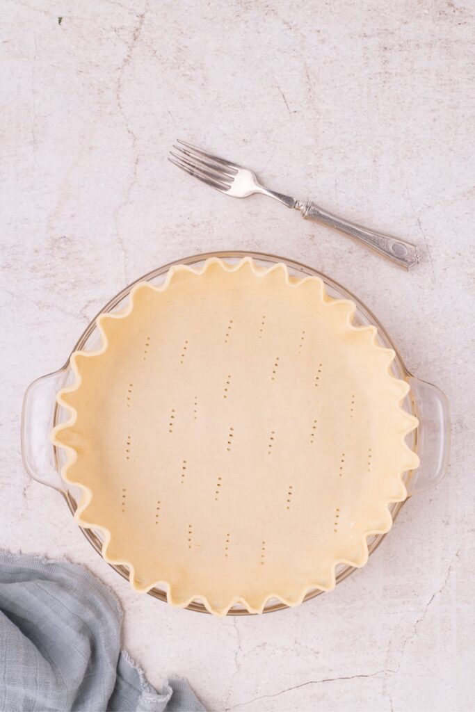 Unbaked pie shell lined in a glass pie plate. The pie shell has been poked several times at the bottom of the shell with a fork. The fork sits beside the pie plate.