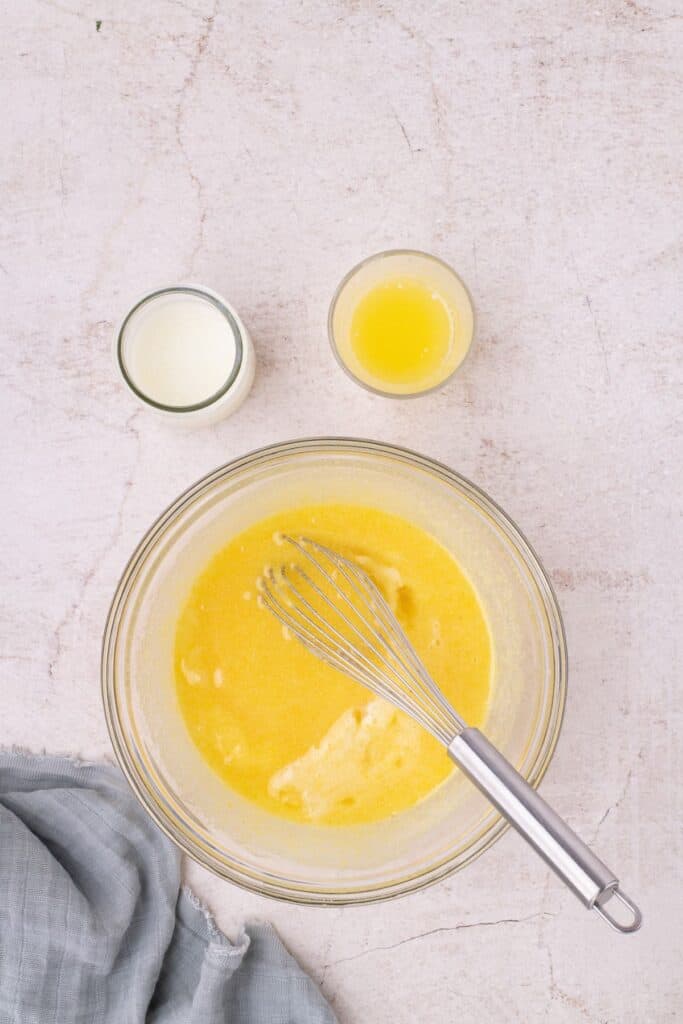 Mixture of chess pie filling is in a glass bowl with a whisk. Small jars of buttermilk and butter sit above the mixing bowl.