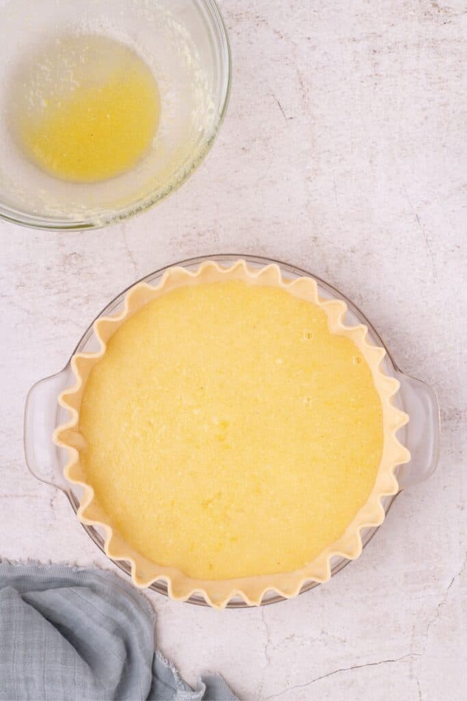 Unbaked mixture for chess pie in an pie shell. An empty glass mixing bowl is above it.