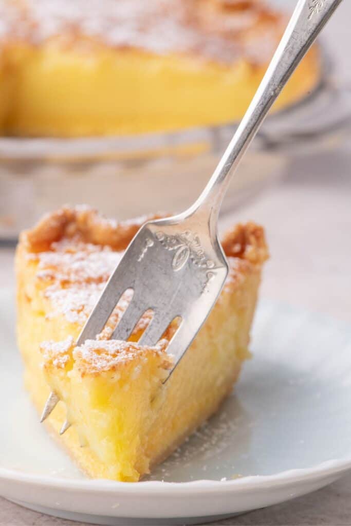 A slice of chess pie is on a white serving plate. A fork is removing a bite from the piece of pie.