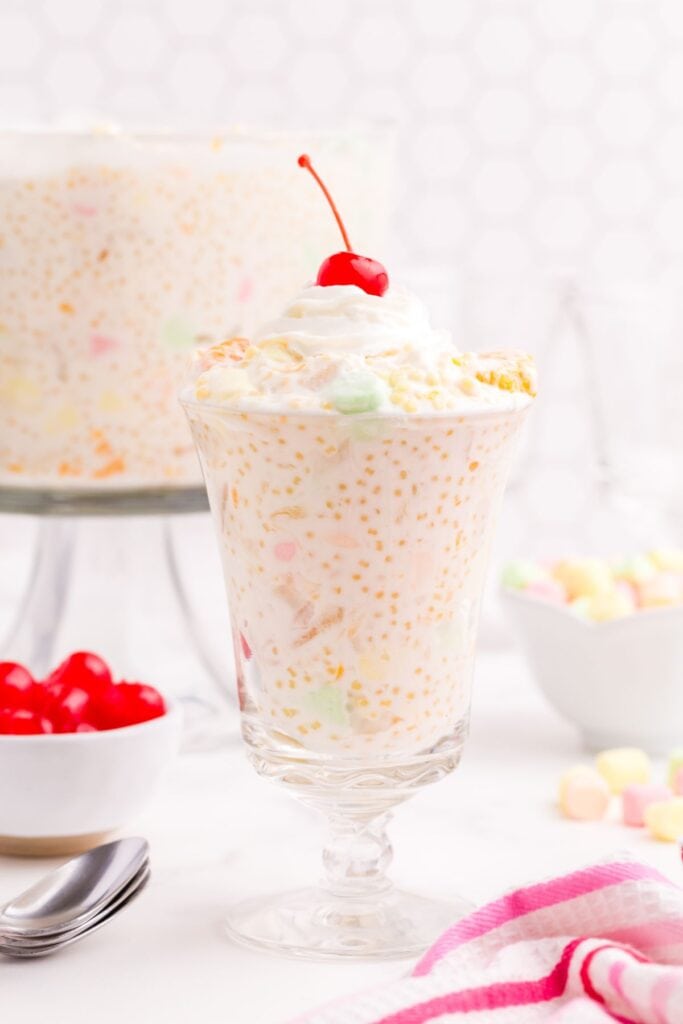 Serving glass of frogs eye salad with whipped cream and a Maraschino cherry on top. There is a large serving bowl of frogs eye salad in the background with small bowls of Maraschino cherries and pastel colored mini marshmallows.