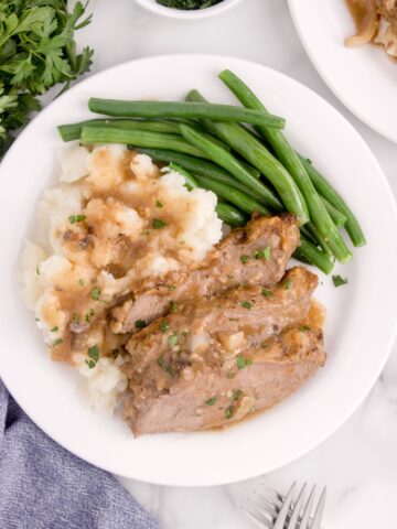 Overhead view of round steak on a white dinner plate with mashed potatoes and green beans. Round steak and mashed potatoes are drizzled in gravy.