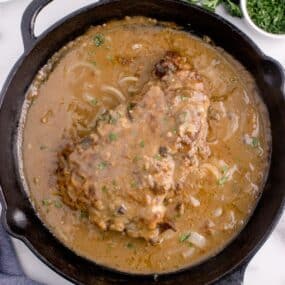 Cast iron pan with completed round steak and gravy. Pan surrounded by a small bowl of parsley, bowl of mashed potatoes, and a white plate of green beans.