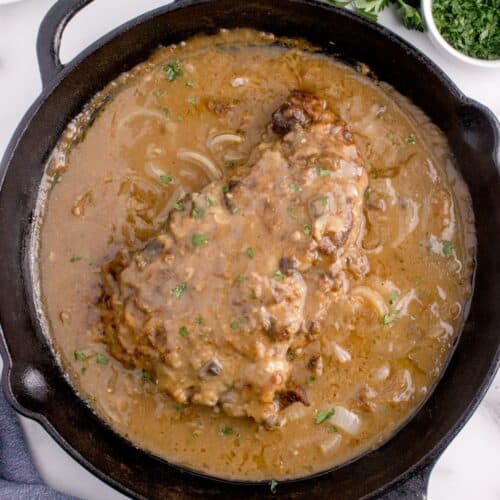 Cast iron pan with completed round steak and gravy. Pan surrounded by a small bowl of parsley, bowl of mashed potatoes, and a white plate of green beans.