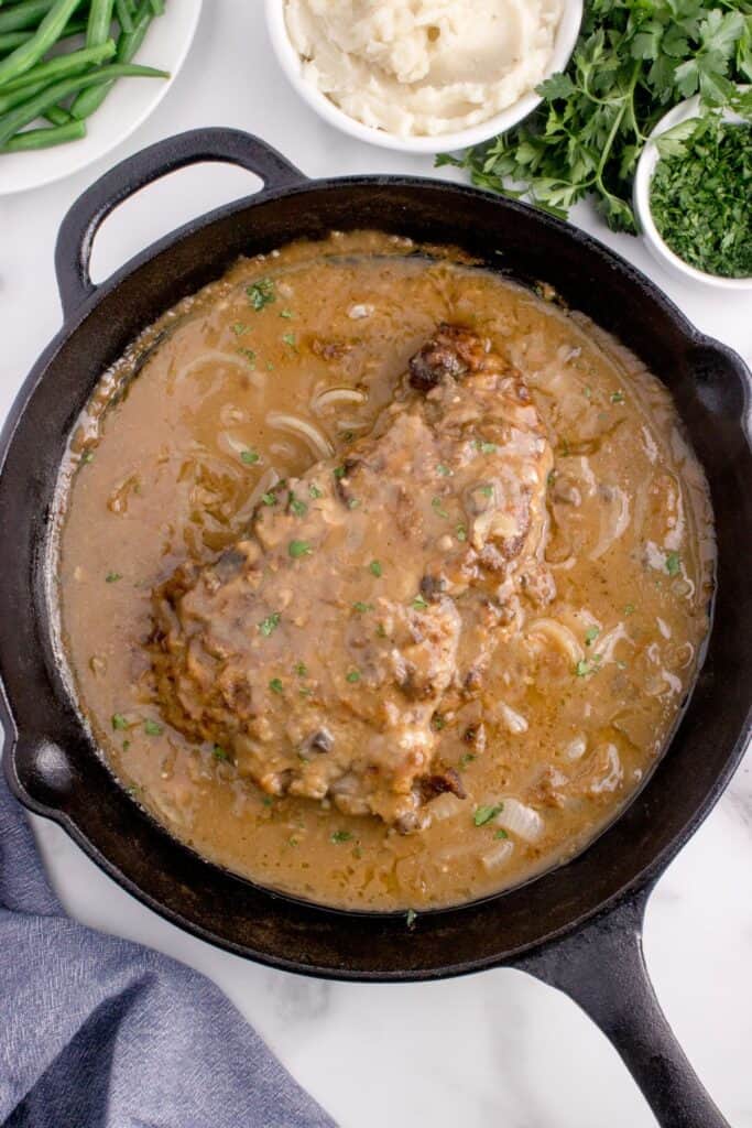 Cast iron pan with completed round steak and gravy. Pan surrounded by a small bowl of parsley, bowl of mashed potatoes, and a white plate of green beans.