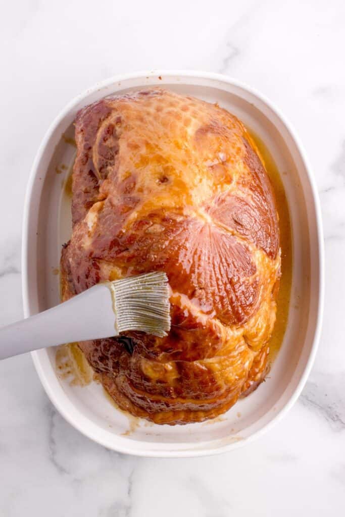 Spiral ham in a white baking dish. A basting brush is covering the ham in a glaze.