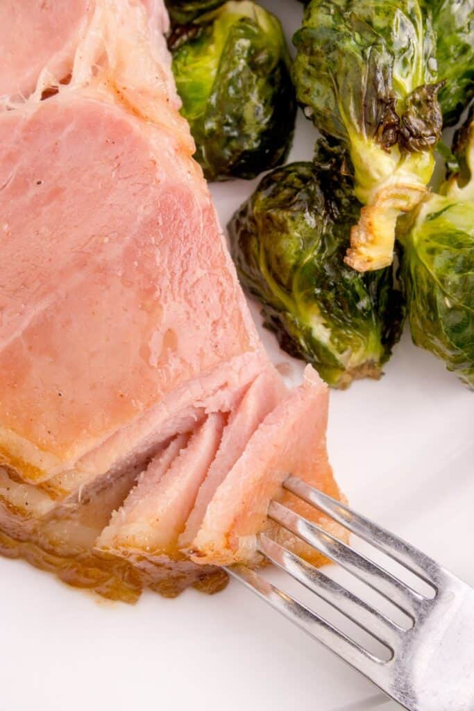 Close up image of several slices of spiral ham and brussel sprouts on a plate. A fork is pulling apart a bite of ham.