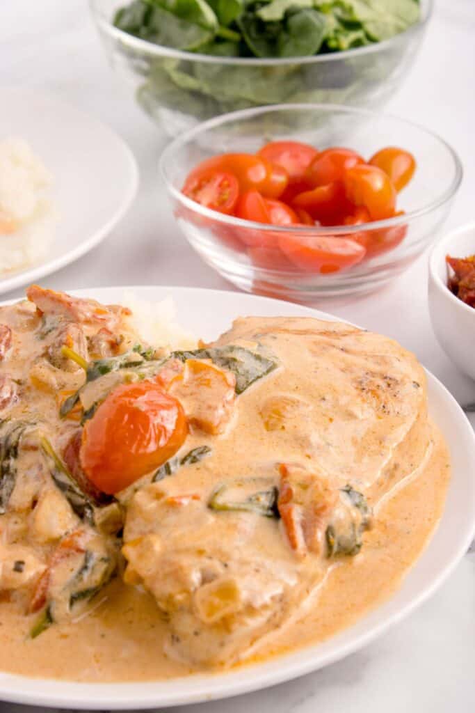 Completed Tuscan Chicken on a white dinner plate with small glass bowls of spinach, tomatoes, and sun dried tomatoes in the background.