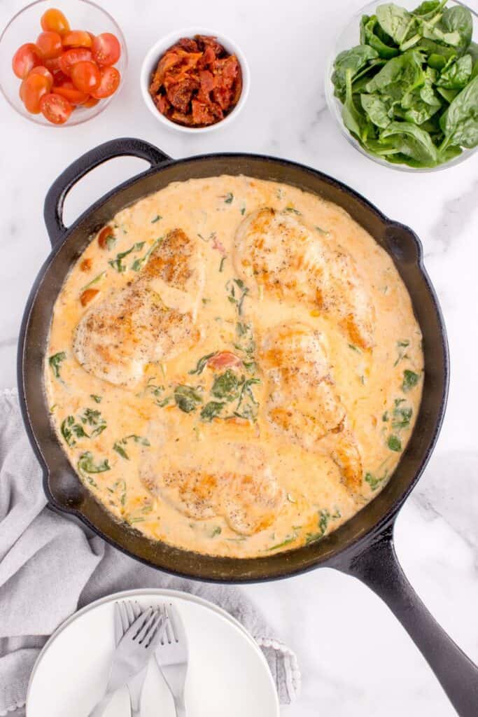 Completed Tuscan chicken in a cast iron skillet. Skillet is surrounded by small bowls of tomatoes, sun dried tomatoes, and spinach.