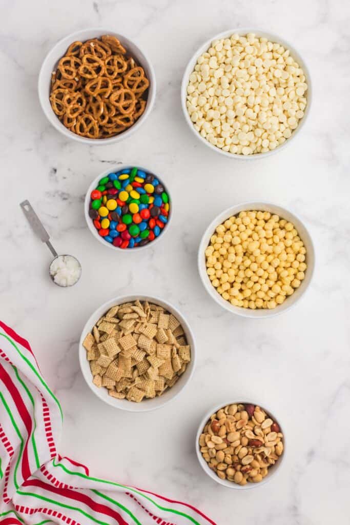 Ingredients for white trash candy (white chocolate chex mix candy): rice chex cereal, kix cereal, mini pretzels, M&Ms, salted peanuts, white chocolate chips, and coconut oil.