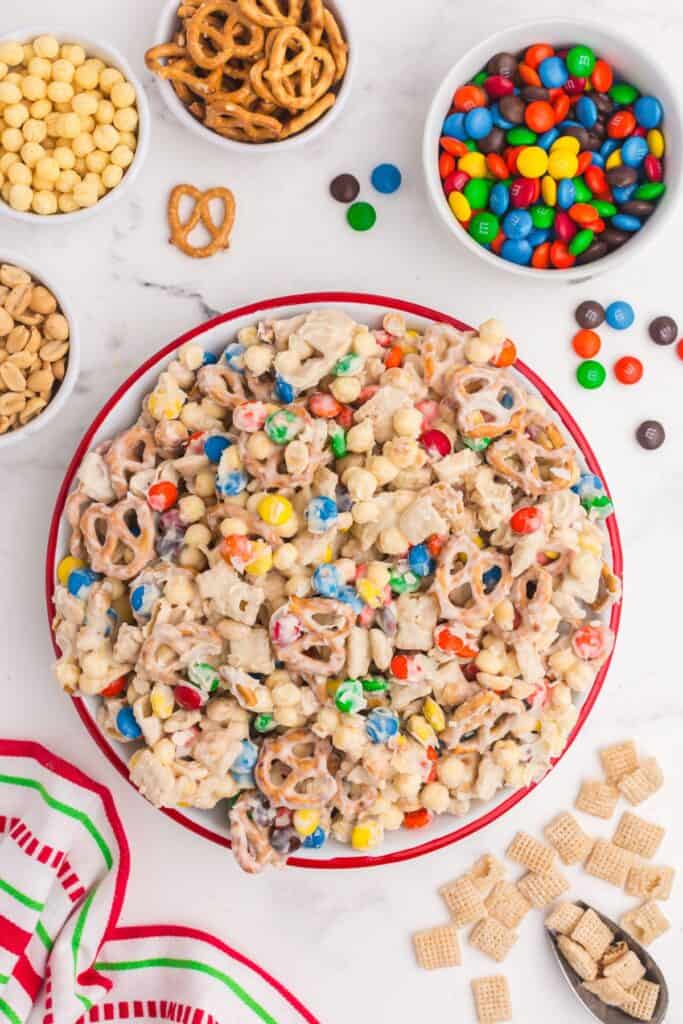 White trash candy (white chocolate chex mix candy) in a serving bowl surrounded by small bowls of peanuts, cereal, pretzels, and Christmas colored M&Ms.